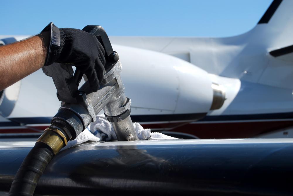 The image shows a cropped photograph of a fuel pump refuelling and aircraft on wing. The Hand holding the pump and pump are left of centre and in sharp focus with the background blurred.