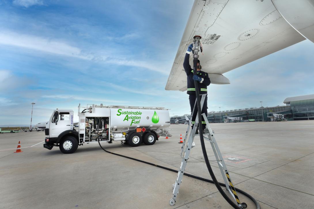 An airport scene showing a worker refuelling an aircraft wing using a hose connected to a fuel truck labelled 'Sustainable Aviation Fuel.' The worker is standing on a ladder, and the clear sky and airport infrastructure are visible in the background.